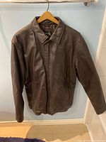 Front view of a men's dark brown genuine leather jacket with zipper closure, hanging on a hanger against a blue wall.