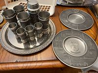 Pewter drinking vessels in set of different sizes stacked on a pewter tray, showing embossed detailed scenes on cups.