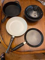 Four pieces of cookware on a wooden table including cast iron skillet, enameled Dutch oven with lid, and two nonstick frying pans with handles