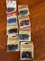 Nine packs of assorted glass beads arranged on wood surface. Includes rust mix, blue mix, amethyst, and other multicolor bead assortments. Various branded packaging visible including Elements and A Touch of Glass.