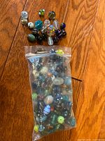 Lot of assorted glass beads inside a clear plastic bag and a small group of beads outside on a wooden floor, showing the variety of colors and shapes.