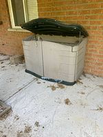 Beige plastic outdoor storage shed with dark green lid and base, placed on concrete beside a brick wall, lid open.
