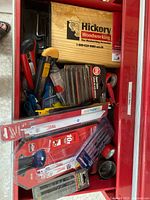 Top view of red tool box drawer containing Hickory wooden box, Milwaukee Sawzall blades, drill bits, pipe wrench, yellow and black handled utility knives, hex keys, and other cutting tools.
