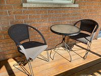 Full view of two black bistro chairs and one round table on wooden deck against brick wall in sunlight