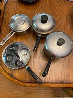 Overview photo of three saucepans with varying lids and an egg poacher insert on a wooden table.