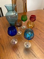 Full view of the large blue-green glass vase and six colored glass goblets arranged on a wooden table.