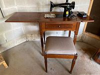 Full view of Singer sewing machine set up in wooden sewing cabinet with matching wooden stool with cushion seat.