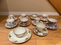 Full view of 8 teacup and saucer sets arranged on hardwood floor showing the variety of floral patterns and gold trim