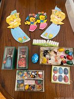 Overview photo showing large paper chick cutouts, boxed nutcracker figures, boxed miniature figures, wooden painted eggs in sets, and small plush chicks with hanging loops.