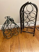 Two ornate metal wine racks on a wooden floor, one green and one black, showing their size and design details.