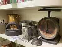 Shelf showing large brass pitcher, cast iron flat iron, steel mixing bowl, two metal graters, silverplate butter dish lid, vintage scale with dial, and small metal cup.