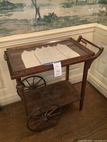 Front view of antique wooden tea cart with large metal wheels, glass top, and lace doily on top shelf.