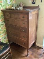 Full view of the antique wooden tall dresser showing six drawers with mismatched brass pulls, some drawer locks visible, placed in corner with floral wallpaper backdrop.