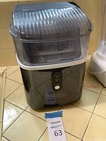 Front and top view of the black Cowsar countertop ice maker with clear plastic top cover, placed on a tile floor.