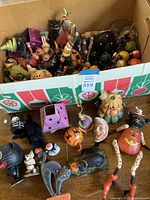 Box of assorted Halloween decorations in cardboard box with green and red graphics and several figurines spilled in front, showing overall lot contents