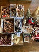 Full view of the assorted vintage Christmas decorations spread out on a table. Includes cloth angel dolls, bottle brush trees, a snowman face decoration, Santa figurine, icicle glass ornaments in box, and various small figurines.