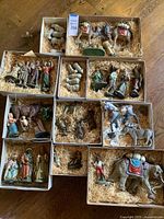 Multiple boxed painted resin or ceramic nativity figurines placed on table with straw filler, showing animals, human figures, and grouped in individual boxes.