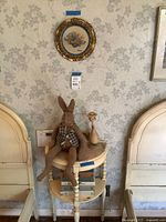 Wide view of cream-painted wooden nightstand between bed headboards, displaying rabbit plush on top, ceramic figurine, and books on lower shelf under floral wall art