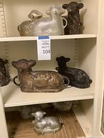 Seven vintage metal lamb cake molds displayed on shelves, showing mixed patina and worn surfaces in silver, dark brown, and rusty colors.
