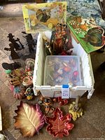 Overview of the lot showing assorted Thanksgiving and autumn themed decorations including ceramic leaves, painted figurines, and a plastic bin with more items.