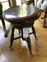 Front angle view of the Victorian-style antique wooden piano stool showing the round seat, turned legs, and metal claw feet on casters with glass balls.
