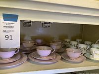 Multiple pink fine china tea cup, saucer, and small plate sets lined up on a shelf, showing cups and bottoms of dishes.
