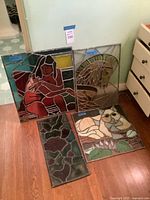 Photo showing all 4 stained glass window panels leaning against a wall on wooden floor. Panels include a woman holding a cat, a horse head, a cat, and an abstract floral pattern.