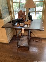 Front view of the wooden side table with turned legs and multiple tiers, placed near a window. Various decorative items seen on top but not included.