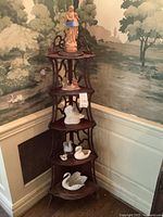 Full view of wooden corner shelf unit in a room corner with five shelves holding ceramic figurines including a woman statue, swans, and birds.