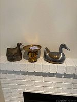 Front view of the three items on a white brick mantle: an antique brass duck figurine on the left, a small brass vase or urn with a flat round removable top in the center, and a silver-colored duck figurine on the right.
