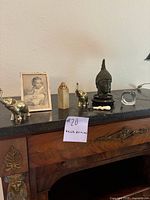 Photo of shelf with antique knick knacks including framed photo, brass elephants, carved stone paperweight, glass sphere, ivory-toned carving, and Buddha head (Buddha excluded from lot).