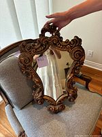 Full view of the antique carved wooden mirror resting on a chair, showing overall shape and carved details.