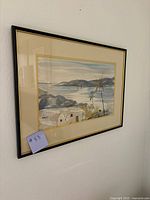 Framed watercolor art piece hanging on a wall, showing the coastal landscape with mountains, water, and buildings.