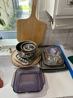 Photo showing wooden paddle cutting board, stainless steel mixing bowls, metal baking sheets, and purple glass Pyrex baking dish on kitchen counter.