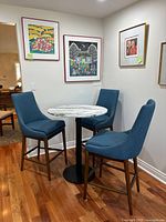 View of round marble patterned table with three blue upholstered barstools around it on hardwood floor, framed wall art on the background wall