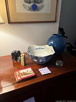 Photo showing all lot items arranged on wood surface: blue vase, white and blue bowl, black elephant figurine, small carved figurine, stack of coasters, and small round tin
