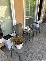 Outdoor patio setup with two metal mesh patio chairs and two glass top metal side tables. One table holds a white ceramic pot with flowers, the other has an empty white pot. Two large white square planters with soil are visible but are not included.
