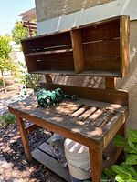 Outdoor wood working table with mounted shelf and green garden hose coiled on top, showing natural wood grain and weathering.