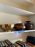 Shelf display of three antique pots/bowls including brass, copper and dark brown pots above books.