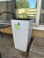 Tall white square tapered garden planter shown on a patio area next to a chair and window.