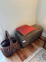 View of woven basket next to rectangular woven storage trunk with red cushion on top.
