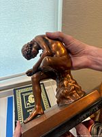 Side view showing the boy sitting on the textured rock base, holding his foot carefully with both hands, close-up of statue detail and patina.