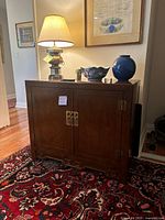 Full front view of vintage wooden cabinet with closed doors, brass hardware, standing on a carpeted floor with decorative items on top.