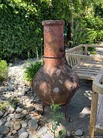 Clay chiminea with a tall chimney and rounded body sitting outdoors on paved ground with garden in background.