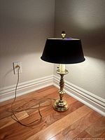 Full view of the vintage brass table lamp with black shade, showing the cord and plug on hardwood floor near a wall outlet.