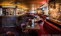 Wide view of the pub interior showing dark wood bar with stained glass details, red tufted bench seating, wooden tables and chairs, and stone walls.