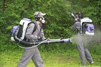 Two operators with Makita backpack sprayers applying tick prevention spray in wooded yard area, wearing protective gear and respirators.