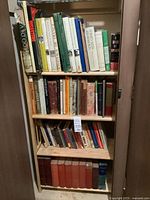 Tall narrow bookcase showing three shelves of assorted books including a bottom row of red clothbound volumes