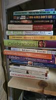 Stack of 14 books including fiction, cookbooks, and self-help on a light wooden chair
