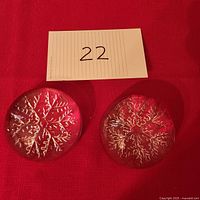 Top view of two round crystal paper weights with visible snowflake designs inside, placed on red fabric background with a numbered tag.